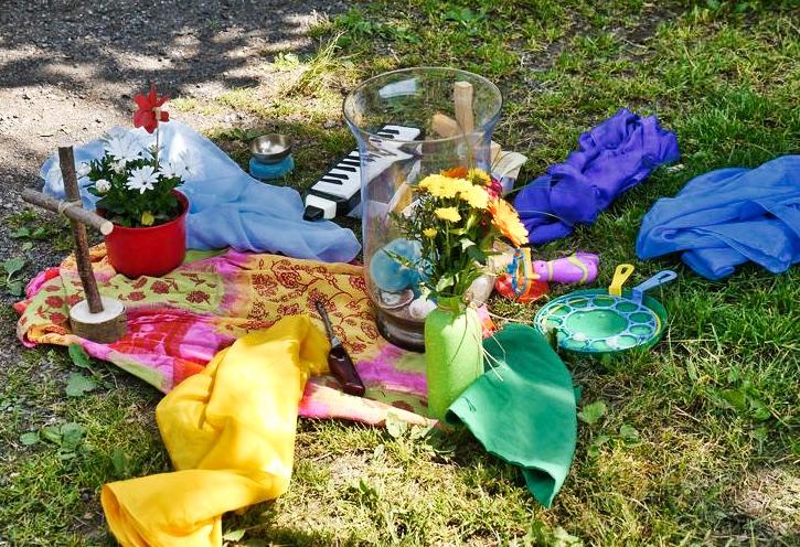 Bunte Tücher auf einer Wiese mit Blumen, Kerzen und einem selbstgemachten Holzkreuz